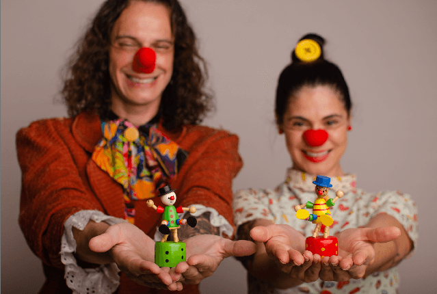 Dois voluntários caracterizados de palhaços, sorrindo e segurando brinquedos de madeira nas mãos